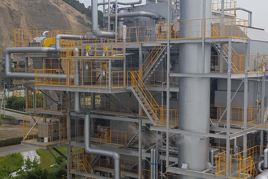 Industrial facility in Kasaoka, Japan, with large metal structures, yellow safety railings, pipelines, and a tall exhaust stack, part of a turnkey greenhouse energy plant.