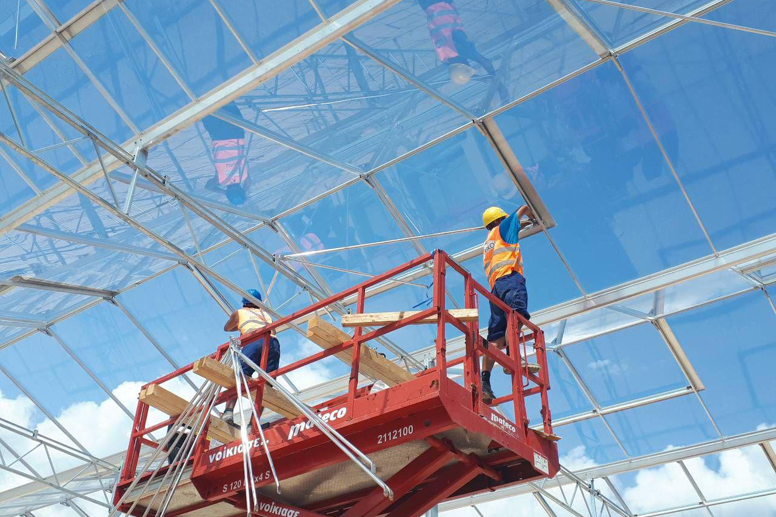 Workers installing greenhouse glass panels while standing on a red aerial lift under a clear blue sky