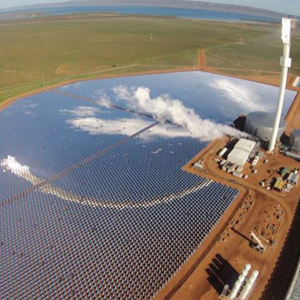 Aerial view of a turnkey greenhouse complex located next to a large solar power plant with a central tower. The facility combines renewable energy with advanced greenhouse technology, showcasing sustainable solutions from expert greenhouse builders.