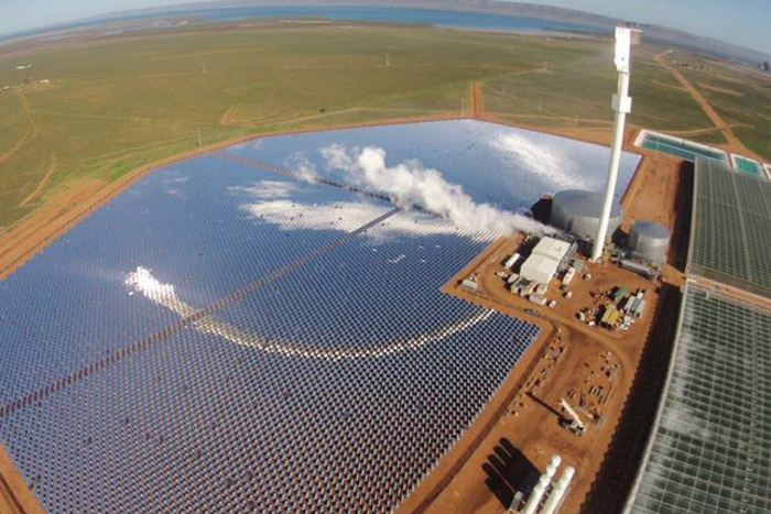 Aerial view of a turnkey greenhouse complex located next to a large solar power plant with a central tower. The facility combines renewable energy with advanced greenhouse technology, showcasing sustainable solutions from expert greenhouse builders.