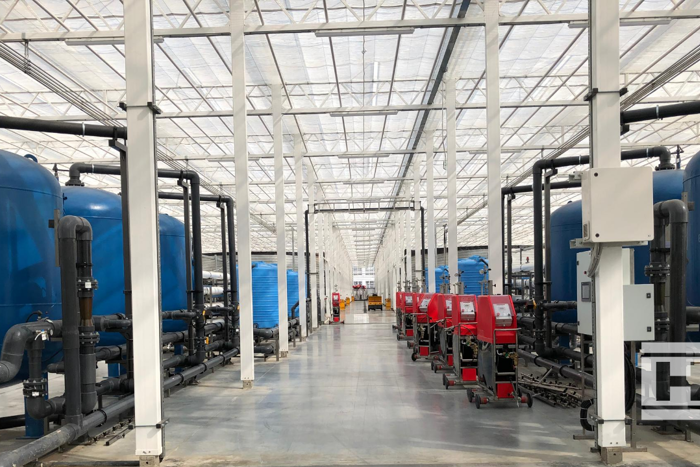 Technical corridor inside Project Norby greenhouse with large blue water tanks, red machinery, and overhead glass roof.