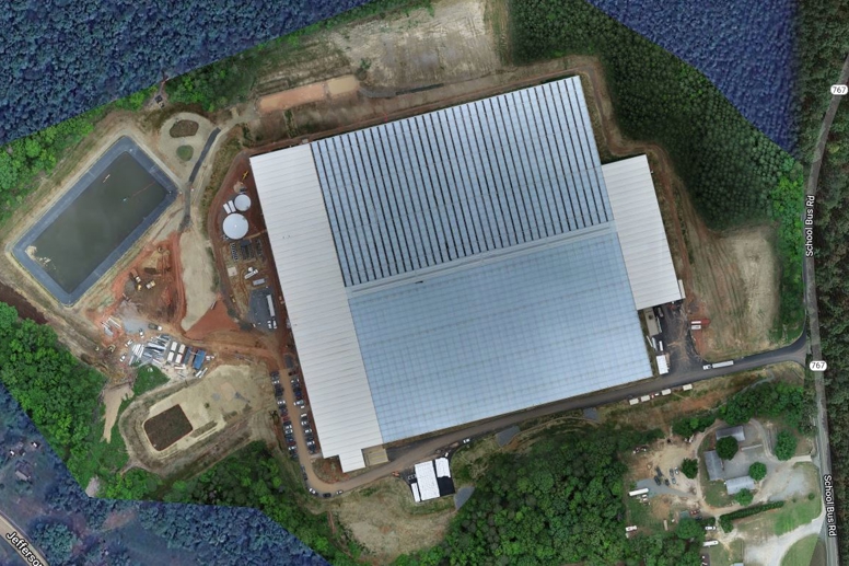 Aerial view of Van der Hoeven’s Better Future Farms greenhouse complex, showing large glass-roofed structure, surrounding forest, water reservoir, and construction site.