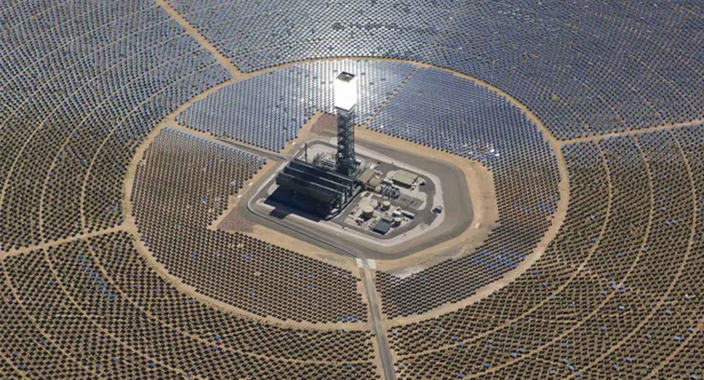 Aerial view of a large solar power plant with a central tower, surrounded by hundreds of mirrored panels concentrating sunlight to generate renewable energy.