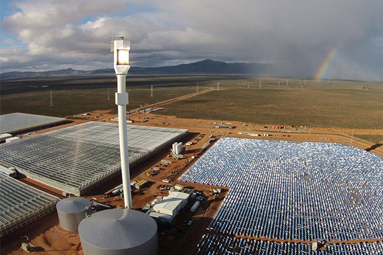 Aerial view of a solar power facility with large reflective panels, water reservoirs, and a tall central tower in a vast desert landscape under a partly cloudy sky.
