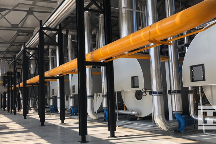 Industrial heating system inside Project Norby greenhouse with large silver boilers, bright yellow pipes, and metal support structures.