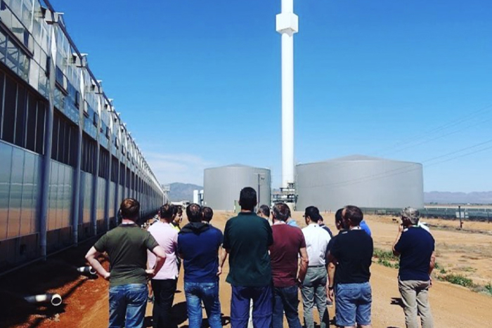 Group of people visiting a greenhouse facility, standing on a dirt road next to the greenhouse building under a clear blue sky