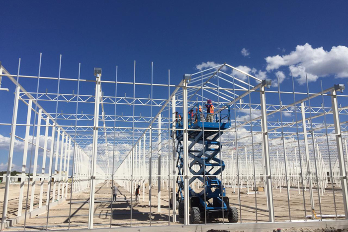 Workers assembling a greenhouse steel structure using a blue scissor lift under a bright blue sky with clouds